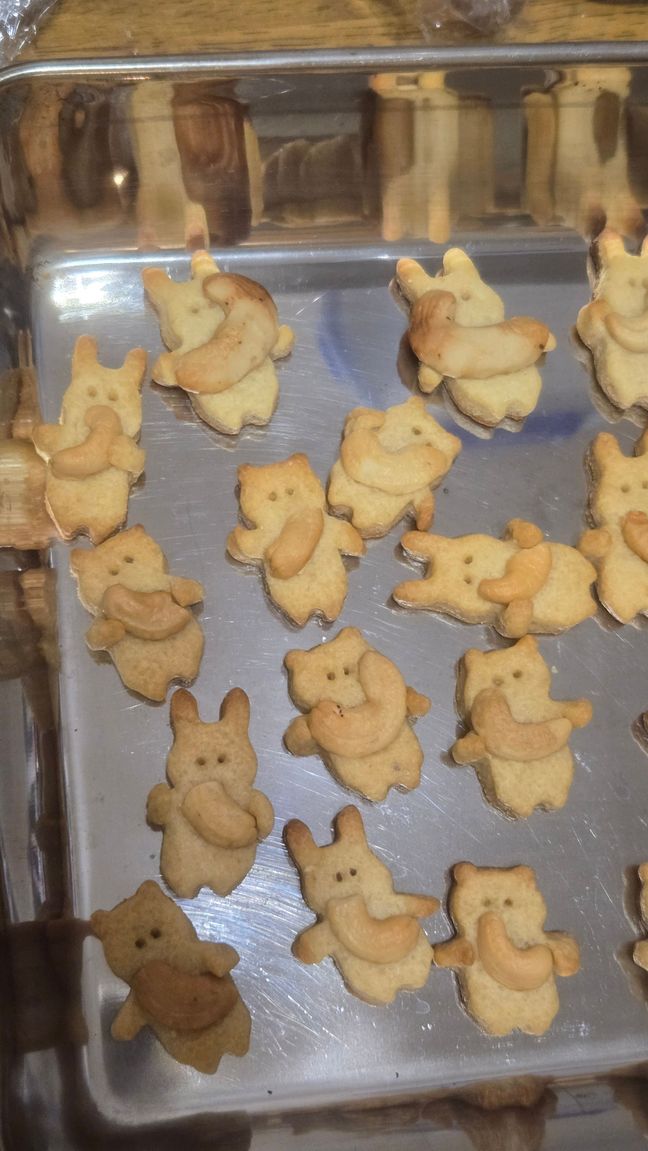 Cute cookies in the shape of a bear and a rabbit. They're lying on a metal baking tray and each hugging a cashew nut.